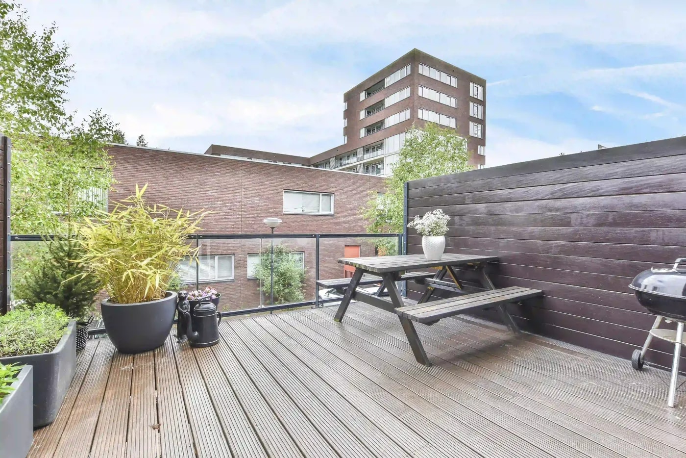 Blick auf eine Dachterrasse mit Holzfußboden, Picknicktisch und Grill, sowie mehreren Grünpflanzen, eines Glasplatten-Geländers und einer Begrenzung aus Holzpanelen.