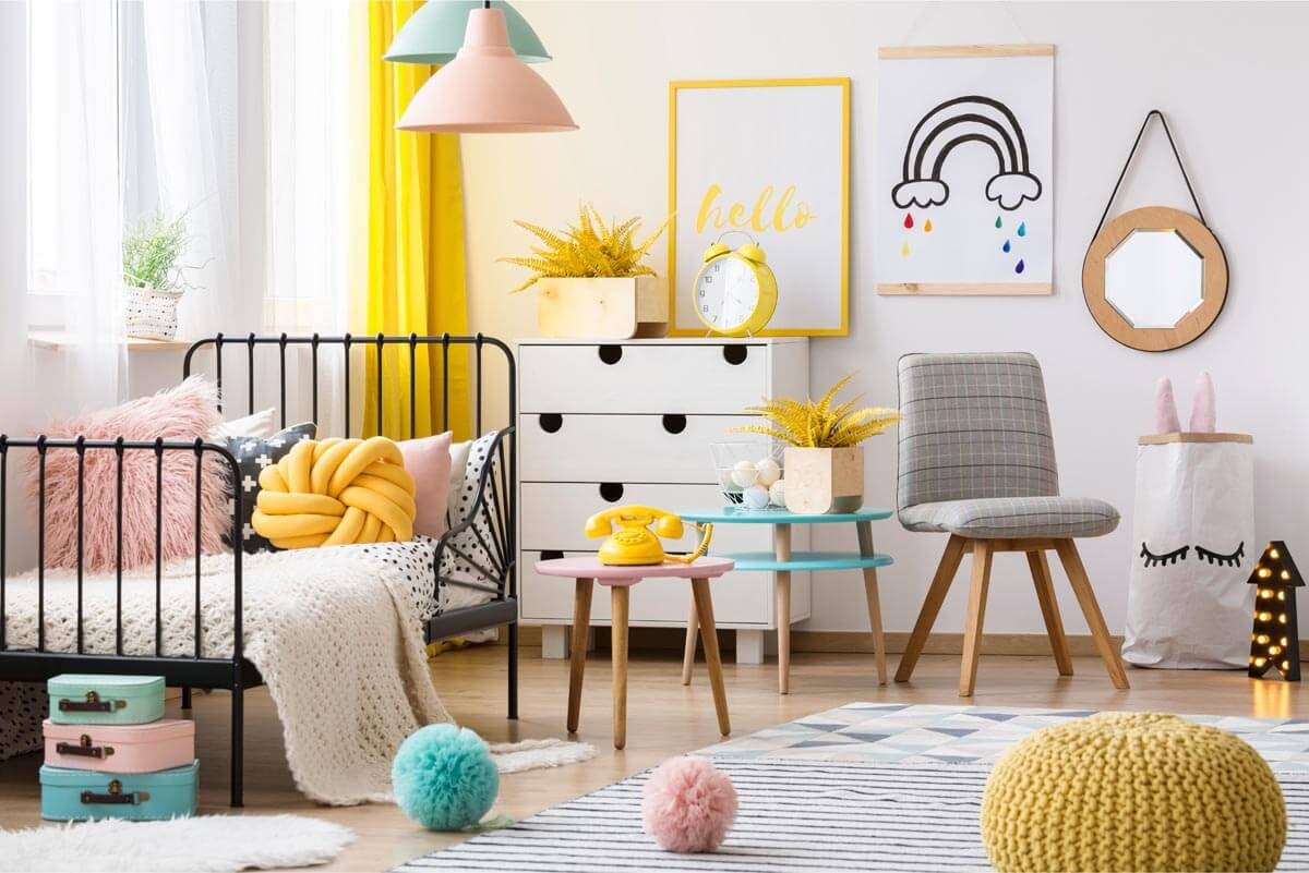 Helles Kinderzimmer mit Fenster und Bett links, vor Kopf ein Sideboard mit Stuhl und Regenbogenbild an der Wand und rechts ein Teppich auf dem verschiedene Plüschbälle liegen.