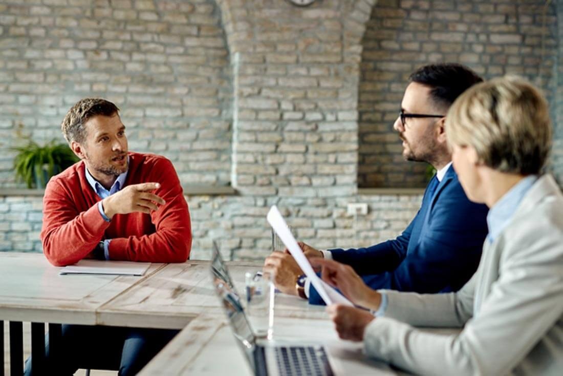 Ein Mann in einem roten Pullover sitzt mit einem Verkäufer und einem Makler an einem Konferenztisch und erläutert etwas, nebem dem zweiten Maklwer steht ein Laptop offen auf dem Tisch.