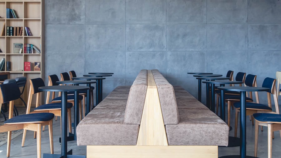 Blick von der Seite auf eine beige Bankgruppe, Rückseite an Rückseite, mit schwarzen Kantinentischen davor und jeweils einem Stuhl vor dem Tisch.