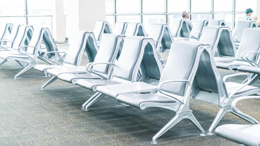 Schräger Blick auf Sitzreihen von metallenen Sitzen in einem typischen Flughafen-Terminal mit breiter Fensterfront im Hintergrund.