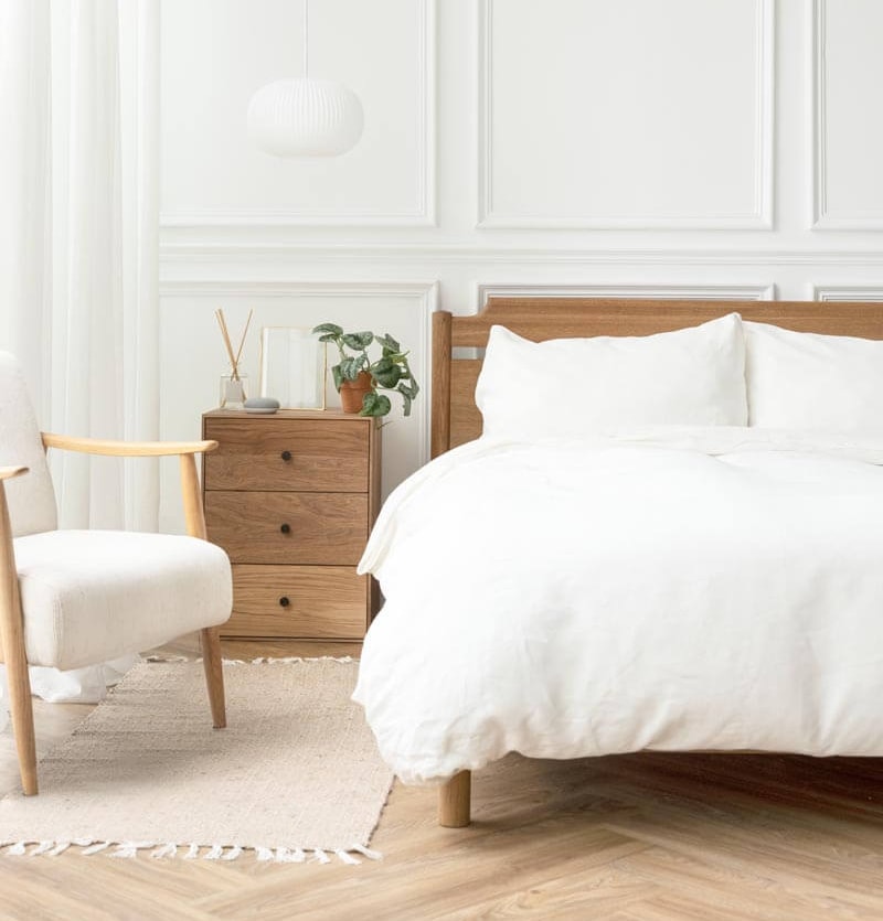 Minimalistisches weißes Schlafzimmer mit Holzbettrahmen, weißer Bettwäsche, Holznachttisch mit Zimmerpflanze, weißem Polstersessel und dekorativen Wandpaneelen.