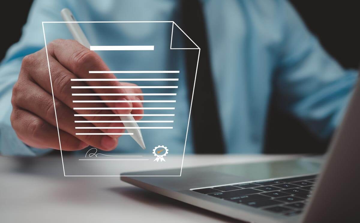 Hand eines Mannes in blauem Hemd hält einen Stift über einem digital schwebend eingeblendeten Vertragsdokument mit Siegel daneben ein aufgeklappter Laptop.