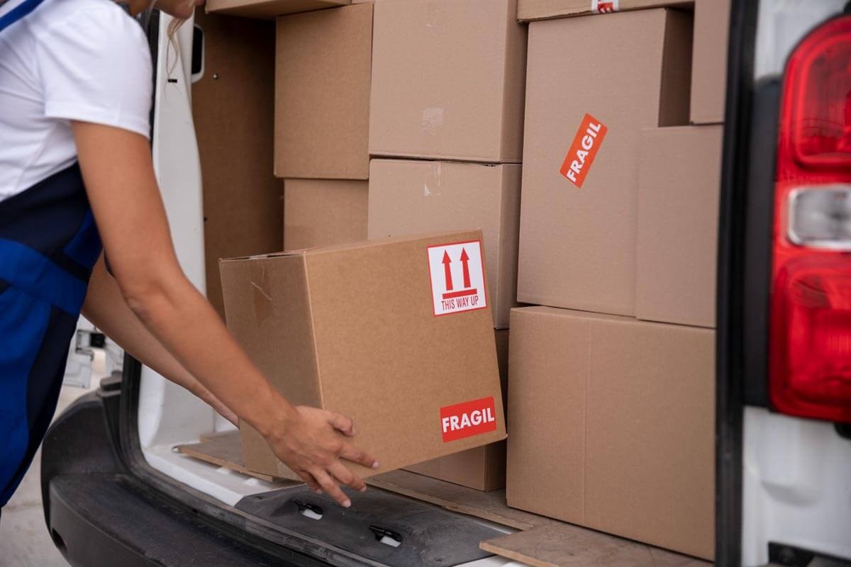 Nahaufnahme: Person in blauer Arbeitskleidung entnimmt einen mit Fragil-Aufkleber beschrifteten Karton aus einem Transporter; im Hintergrund weitere gestapelte Kartons im Laderaum.