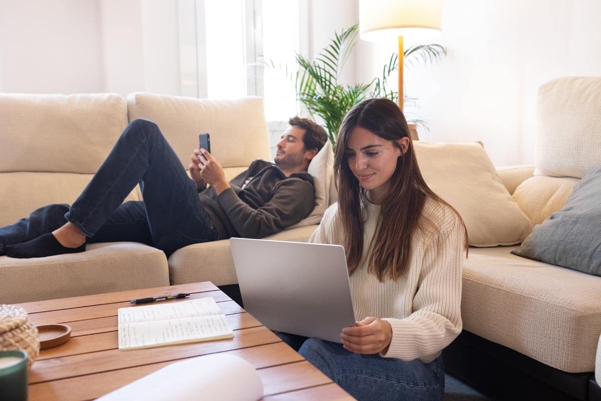 Frau sitzt auf dem Boden zwischen Sofa und einem Couchtisch und arbeitet am Laptop, vor ihr liegen Notizbuch und Stift, im Hintergrund liegt ein Mann entspannt auf dem Sofa und schaut auf sein Smartphone.