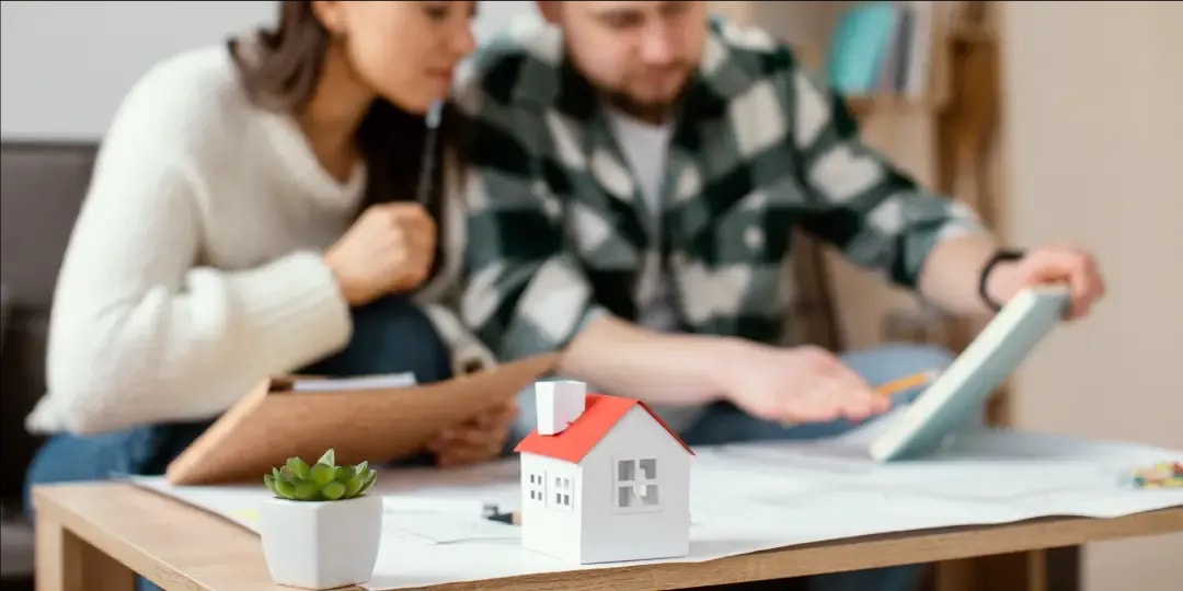 Nahaufnahme einer kleinen Hausminiatur und einer Sukkulente, die die Kante eines Bauplans auf einem Tisch gerade halten, während im Hintergrund, das Pärchen, das vor dem Plan auf der Couch sitzt, über ein Dokument nachdenken.