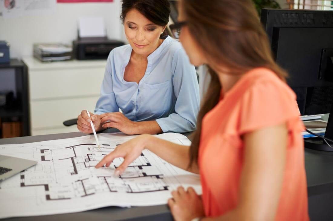 Zwei Frauen sitzen sich an einem Schreibtisch gegenüber und erörtern einen zwischen ihnen auf dem Tisch liegenden Bauplans, jeweils auf ein Detail deutend.