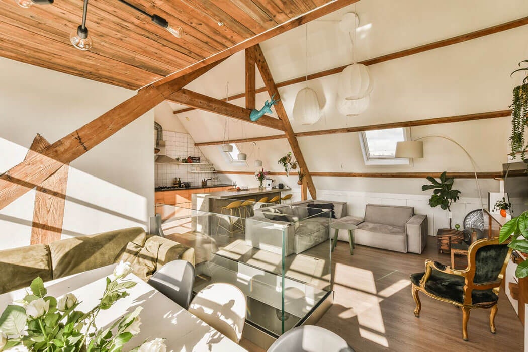 Offen gestaltete Dachgeschosswohnung mit freiliegenden Holzbalken; links ein Esstisch mit weißen Stühlen und Blumenstrauß, mittig eine Glasbrüstung, im Hintergrund offene Küche mit orangefarbenen Fronten, rechts Sofas und ein vergoldeter Sessel mit grünem Samtpolster.