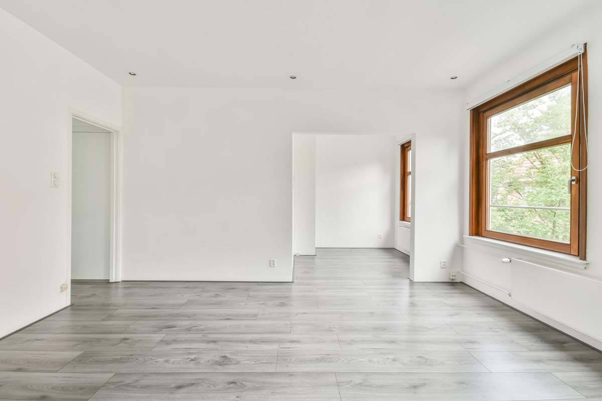 Leerer, heller Raum mit Durchgängen zu Nebenräumen und großem Fenster, mit weißer Wand und hellgrau gemasertem Holzfußboden