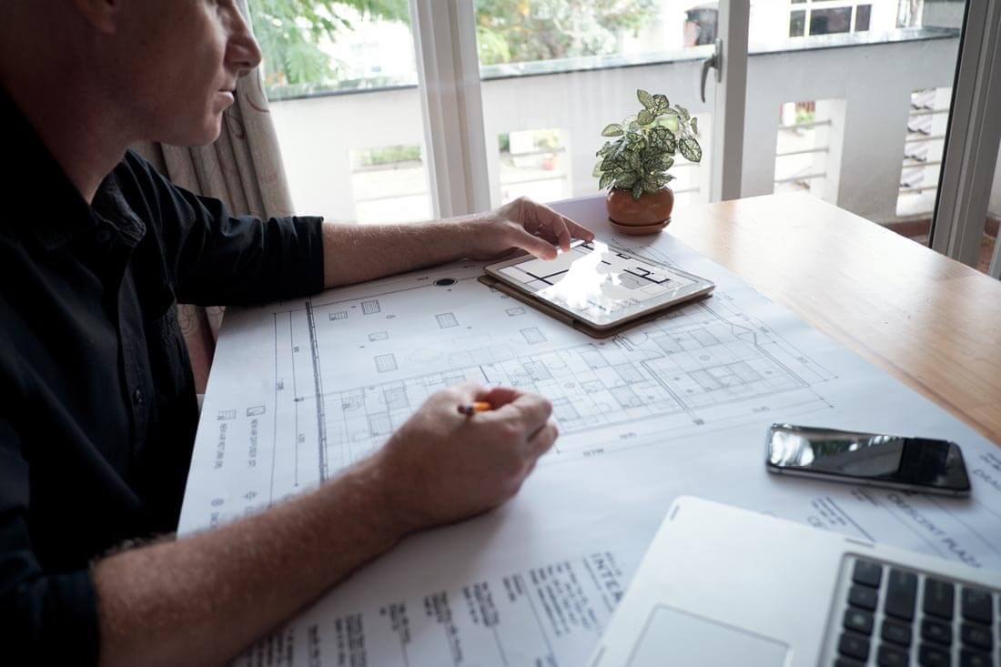 Ein Mann sitzt an einem Holztisch und vergleicht einen vor ihm liegenden Bauplan mit einem Grundriss, der auf einem Tablet an der Seite zu sehen ist.