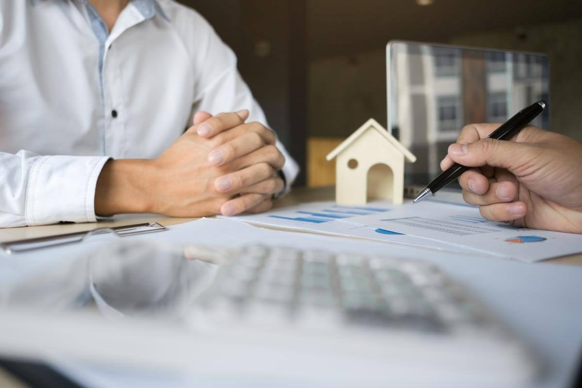 Finanzexperte sitzt am Schreibtisch einer anderen Person gegenüber und erläutert ihr eine Grafik, die zwischen ihnen liegt und und auf deren Details er mit einem Kugelschreiber zeigt.
