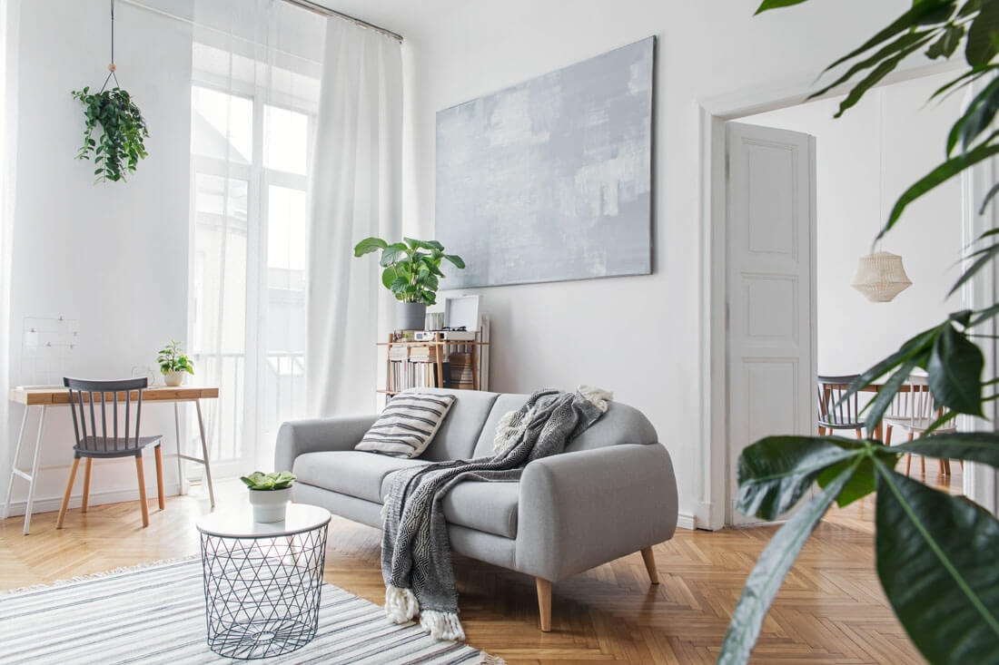 Helles, skandinavisch eingerichtetes Wohnzimmer mit grauem Sofa, gestrickter Decke, Drahtcouchtisch, großer abstrakter Leinwand und Zimmerpflanzen auf Fischgrätparkett.