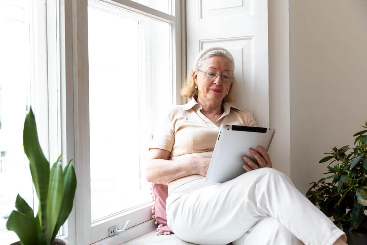 Seniorin sitzt mit Tablet in der Hand und übergeschlagenem Bein auf einer Fensterbank und liest in dem Tablet.