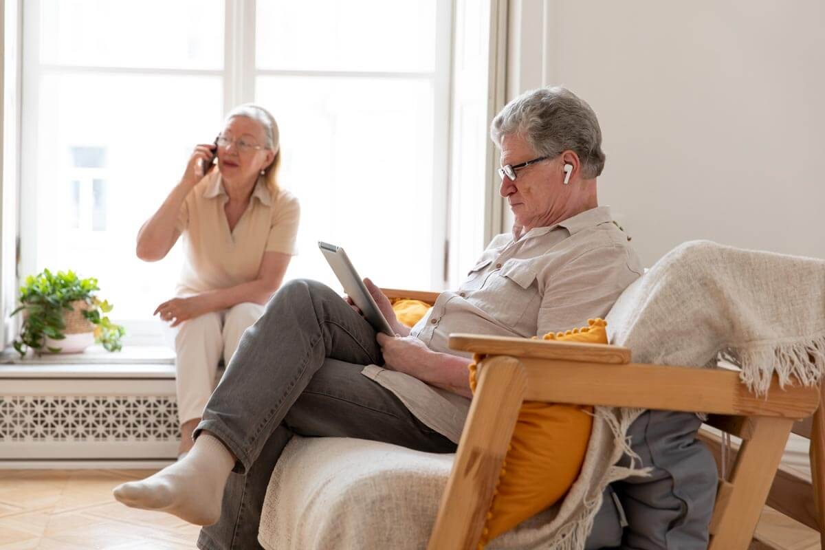 Zwei Senioren sitzen in ihrem Wohnzimmer vorn ein Mann lesend auf der Couch, im Hintergrund eine Frau telefonierend auf der Fensterbank.