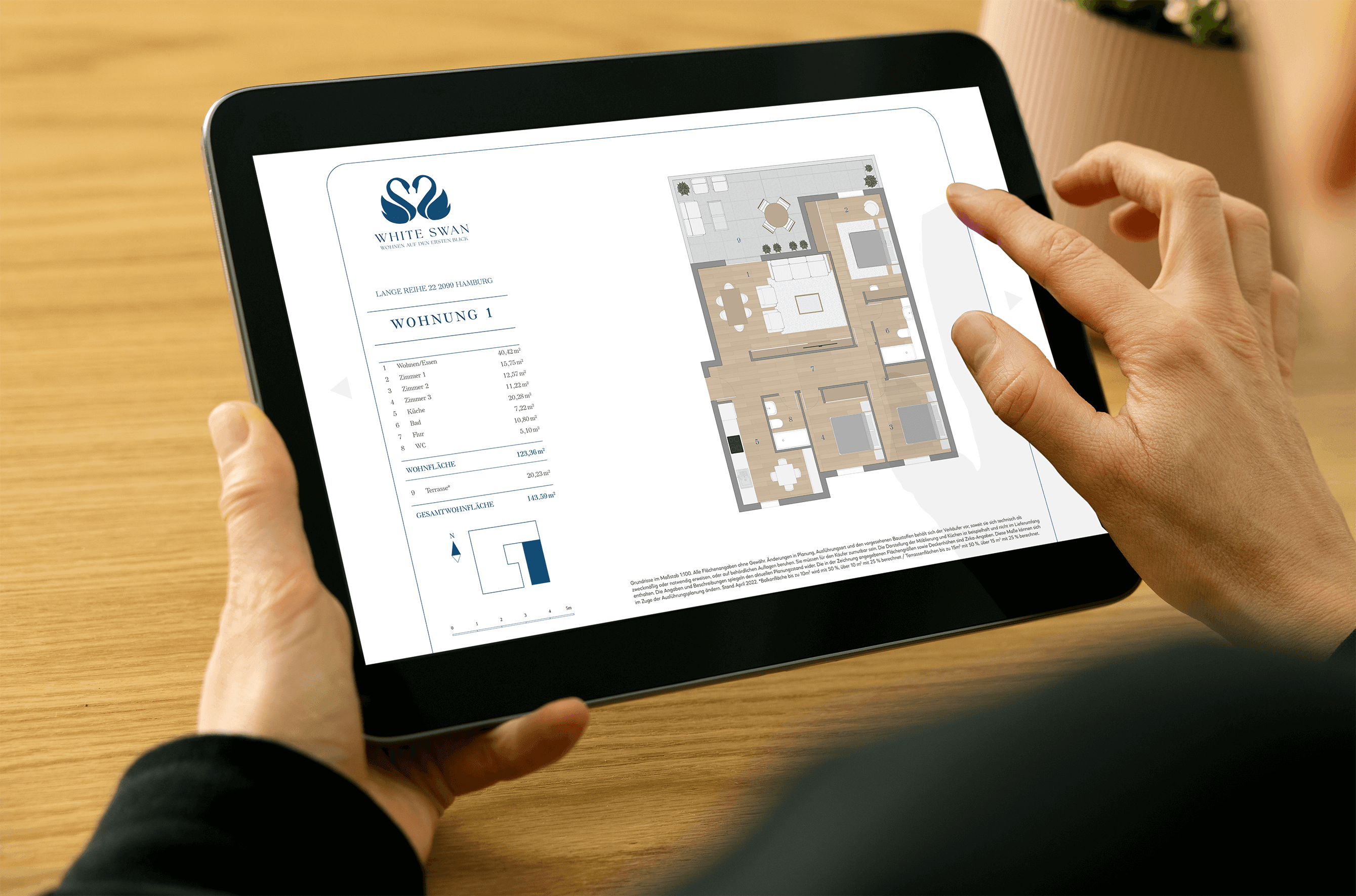 Ein Tablet wird über einem Tisch hochgehalten und ein Zeigefinger swiped gerade eine Exposé-Seite mit einem Grundriss vom Projekt 'Swan Lake' weiter.