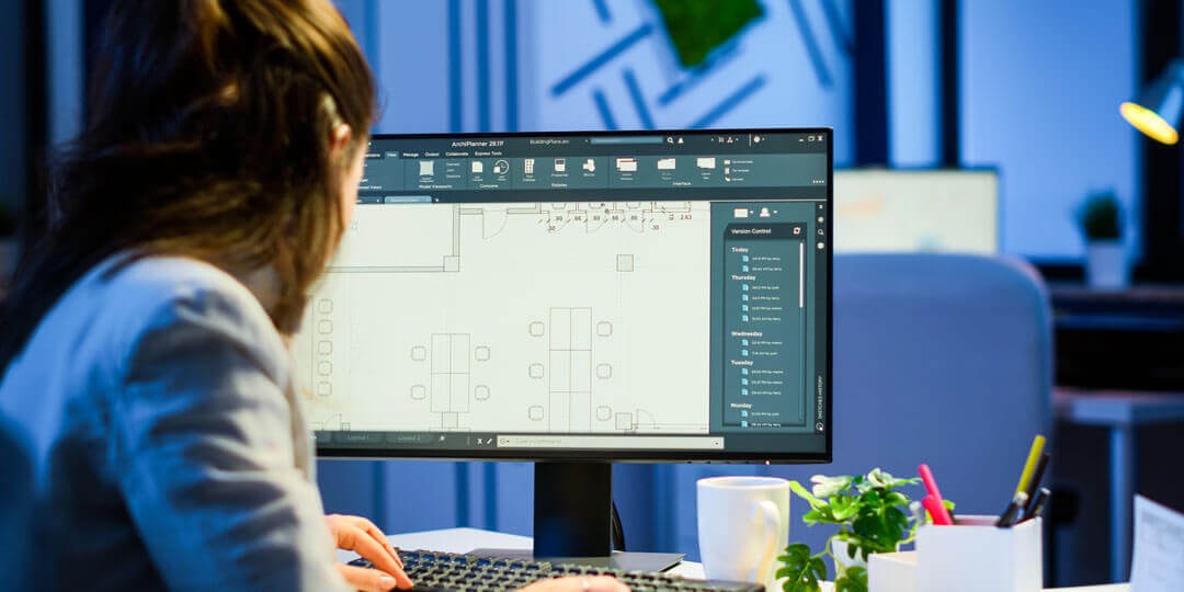 Ansicht von schräg hinten: Frau sitzt vor einem großen MOnitor, der einen Grundriss in AutoCAD zeigt.