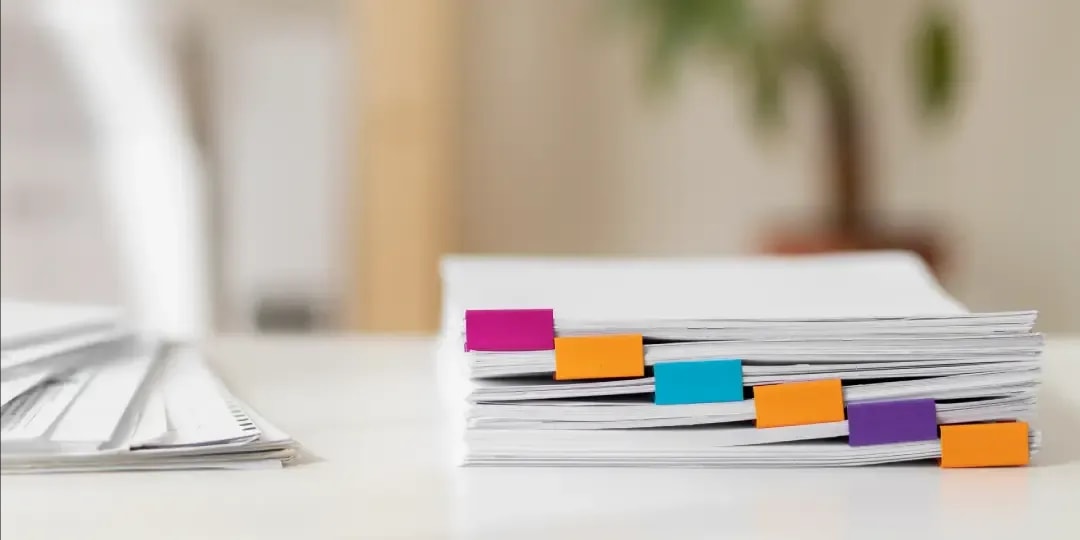Ein Stapel Dokumente auf einem weißen Tisch wobei die einzelnen Dokumente mit bunten Klammer zusammengeheftet sind und daneben liegt ein weiterer Stapel Dokumente.