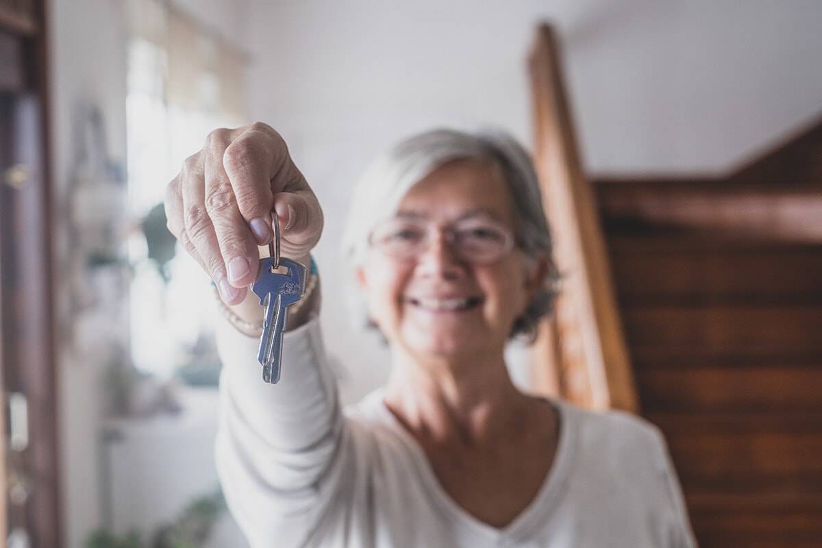 Ältere Dame hält lächelnd in einem Eingangsbereich eines Hauses einen haustürschlüssel hoch.