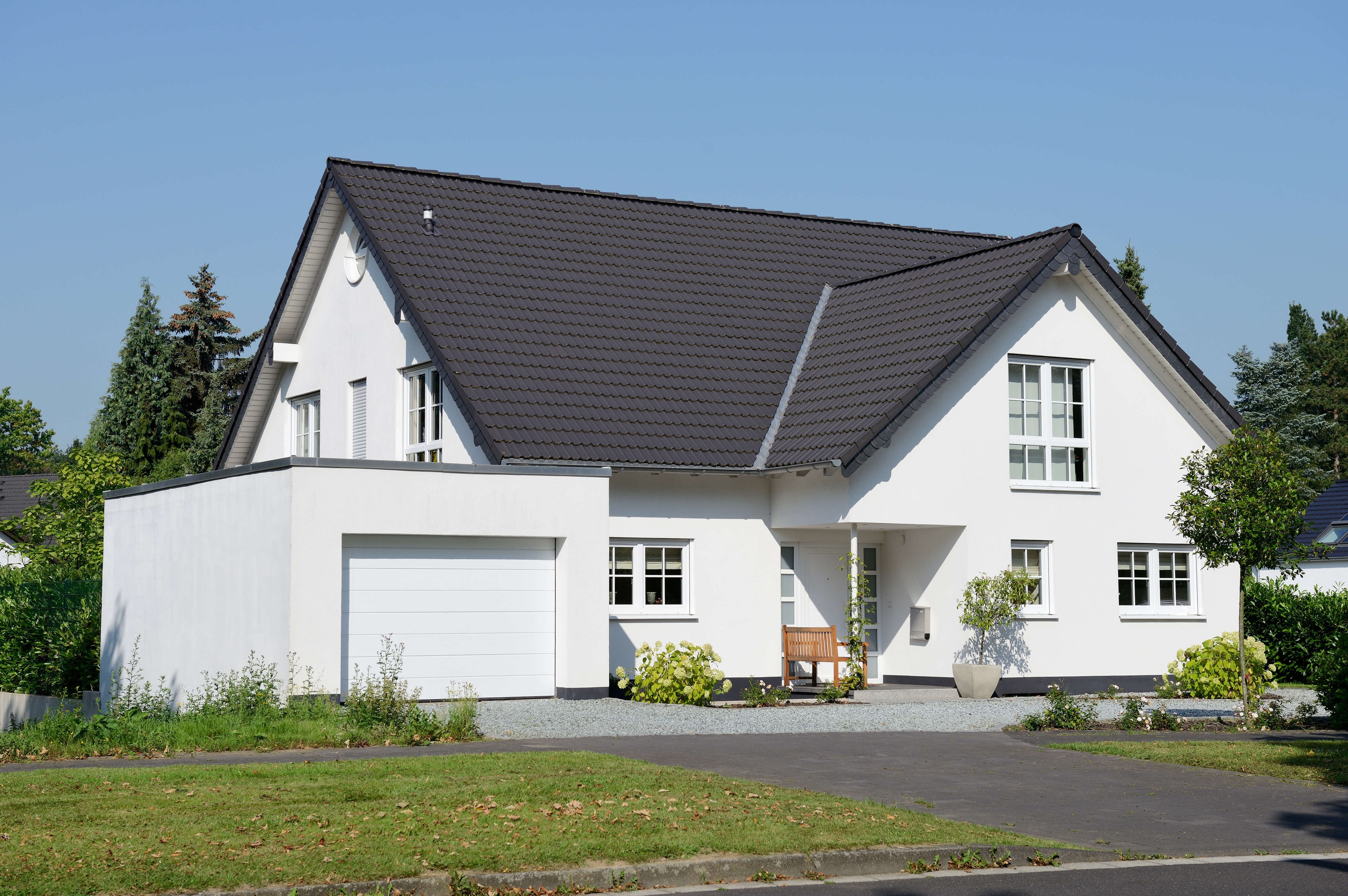 Mehrgeschossiges Einfamilienhaus mit L-Grundform, Satteldach und Flachdach-Garage im Vordergrund, zwischen Grünflächen gelegen. 