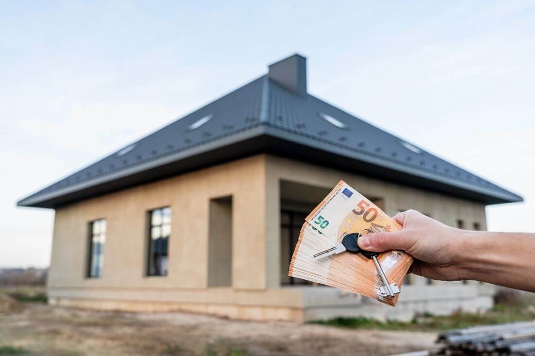 Hand hält Euroscheine und einen Hausschlüssel vor einem Neubau-Bungalow im Hintergrund