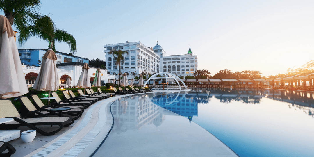 Großzügige Hotelanlage mit weißer Fassade und türkisfarbenem Kuppelturm am frühen Morgen, mit beleuchtetem Außenpool, Liegestühlen und Sonnenschirmen am Poolrand.