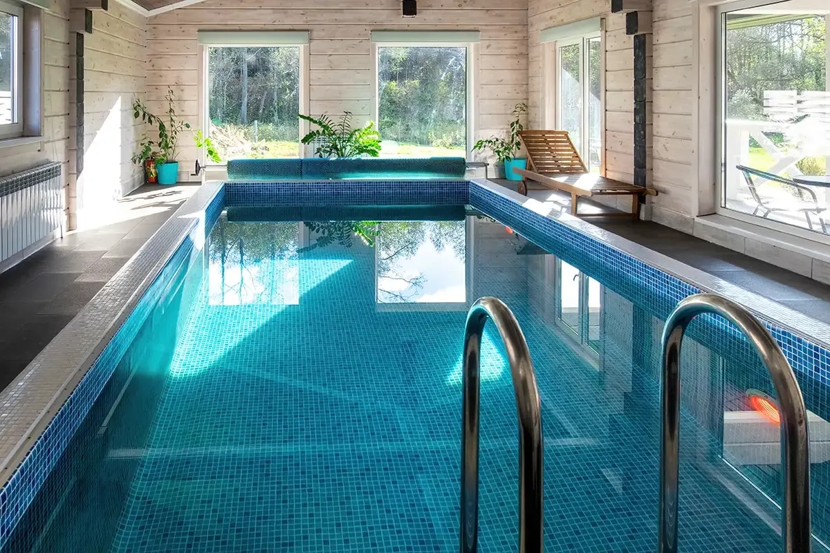Sicht von der kurzen Seite eines Indoor-Pools mit Einstiegsleiter im vorderen Sichtbereich und großen breiten Panorama-Fenstern rundum.