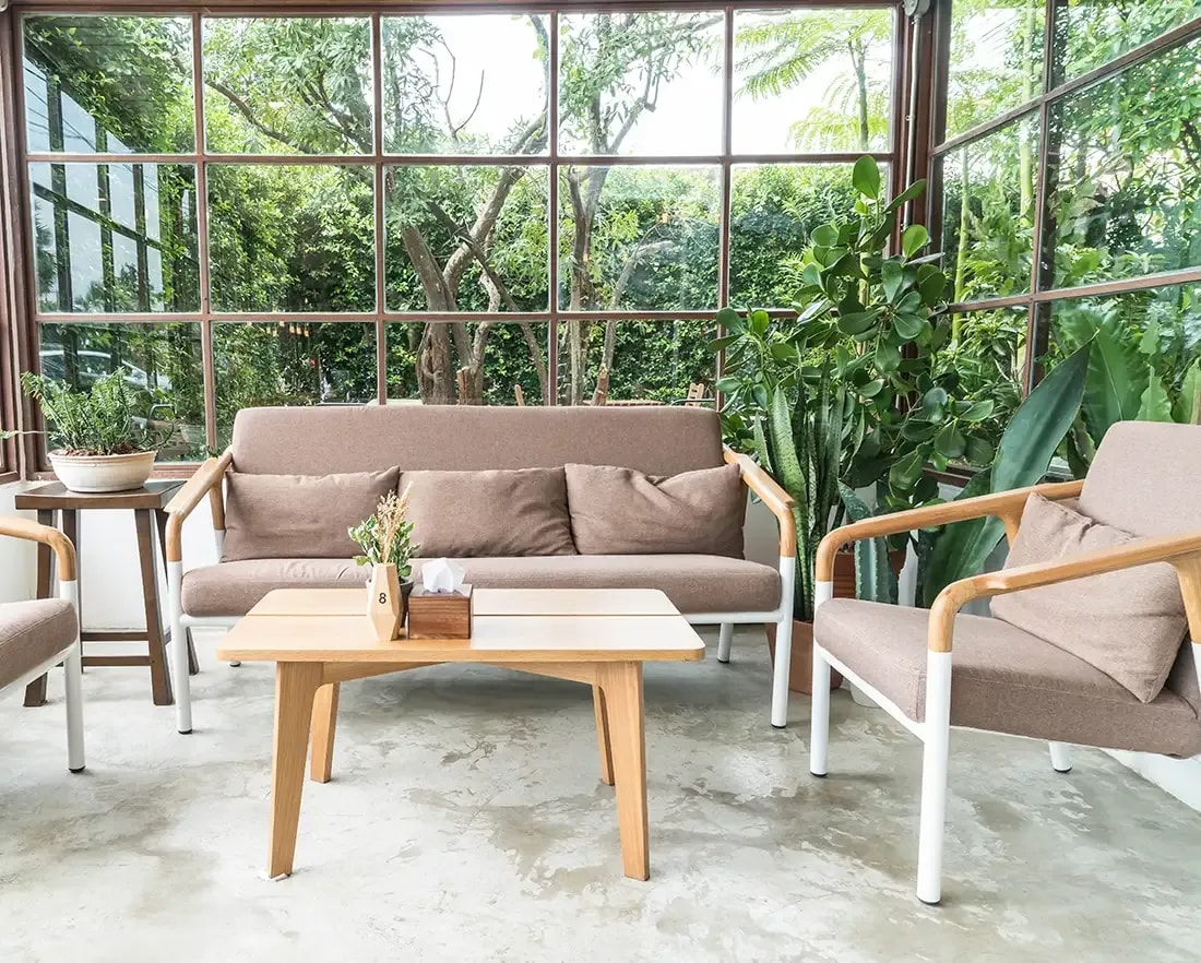 Aufnahme eines Wintergartens mit hell beiger Sitzgruppe vor einer Glasfront durch die üppige Gartenbegrünung erkennbar ist.
