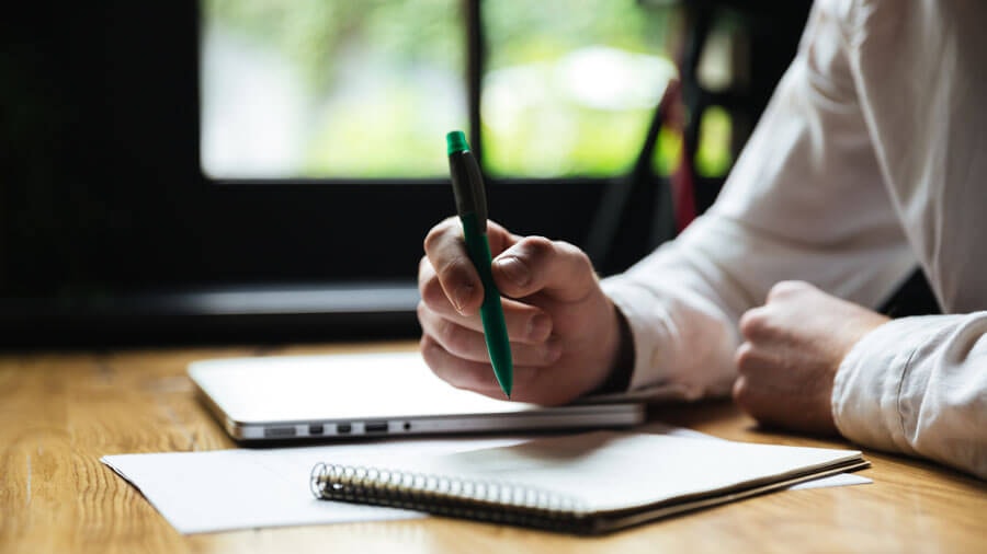 Eine Person sitzt mit einem Stift in der Hand vor einem aufgeschlagenen Notizblock an einem Holzschreibtisch während ein zugeklapptes Laptop daneben liegt.