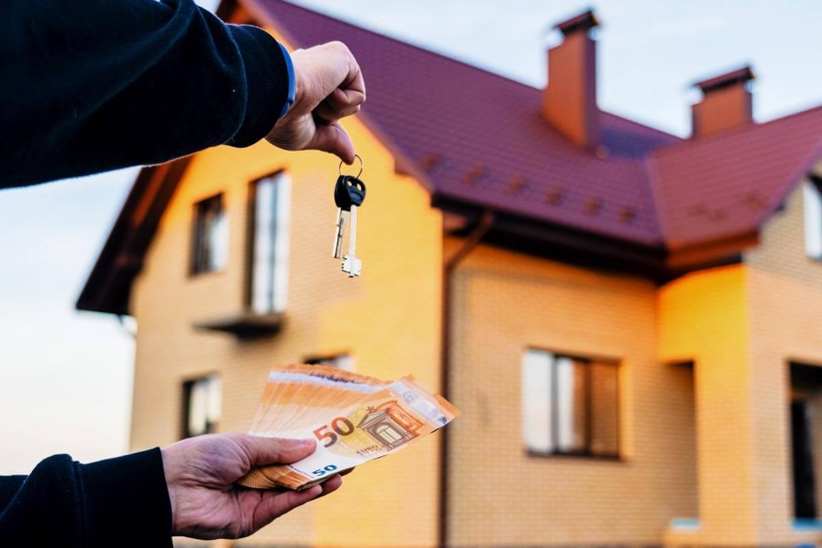 Hände halten einen Hausschlüssel und Geldscheine vor einem Wohngebäude; Symbolbild für Mietkaution und Immobilientransaktion.