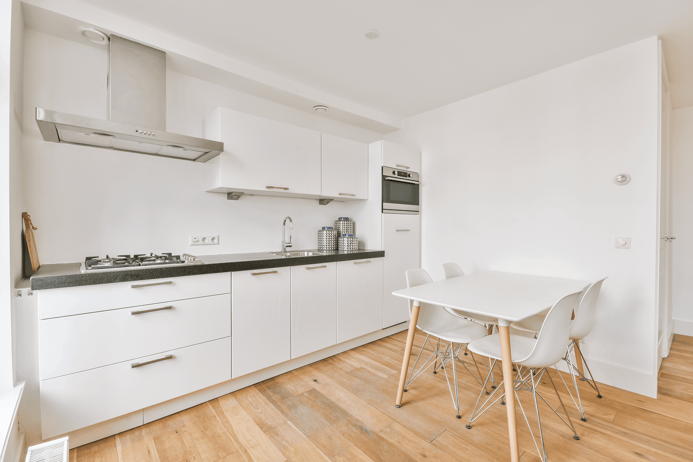 Schlichte weiße Küche mit dunkler Arbeitsplatte, Gasherd und Einbauofen, daneben ein kleiner weißer Esstisch mit Stühlen.