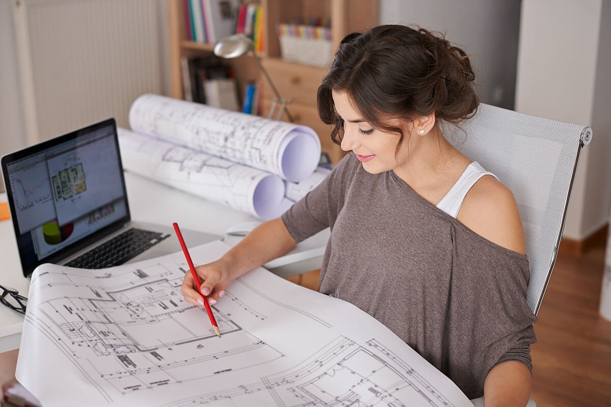 Grafikerin sitzt an einem Schreibtisch mit Laptop und voller Grundrisspläne und notiert Änderungen auf einem aufgerollen Plan vor sich mit einem Bleistift.
