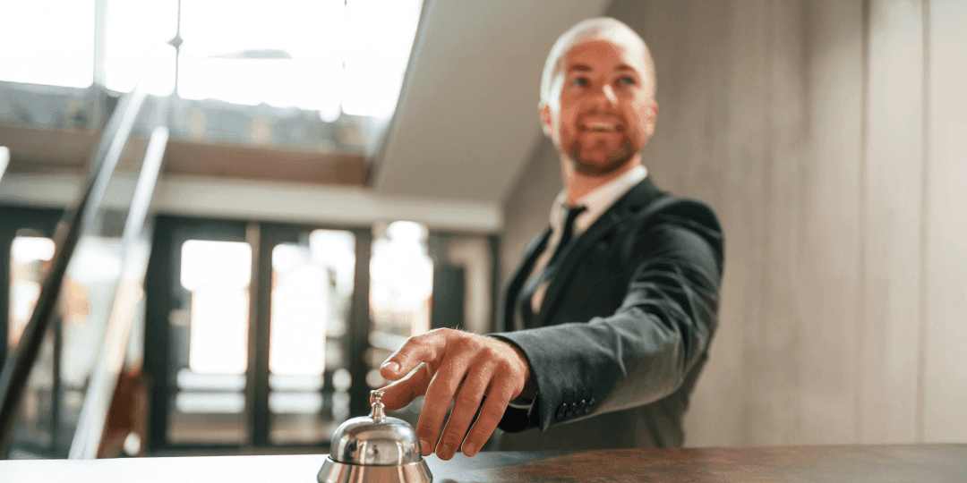 Lächelnder Mann im dunklen Anzug klingelt an der Rezeptionsglocke eines Hotels, im Hintergrund eine helle Hotellobby.
