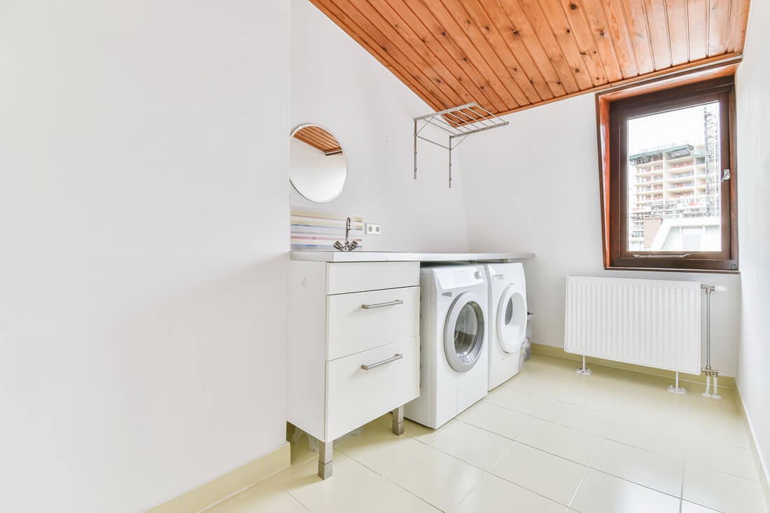 Blick vom Eingangsbereich eines Hauswirtschaftsraumes auf einen Schrank mit Waschbecken, Trocker und eine Waschmaschine mit einem Fenster auf der gegenüberliegende Seite durch das das Nachbarhaus erkennbar ist.
