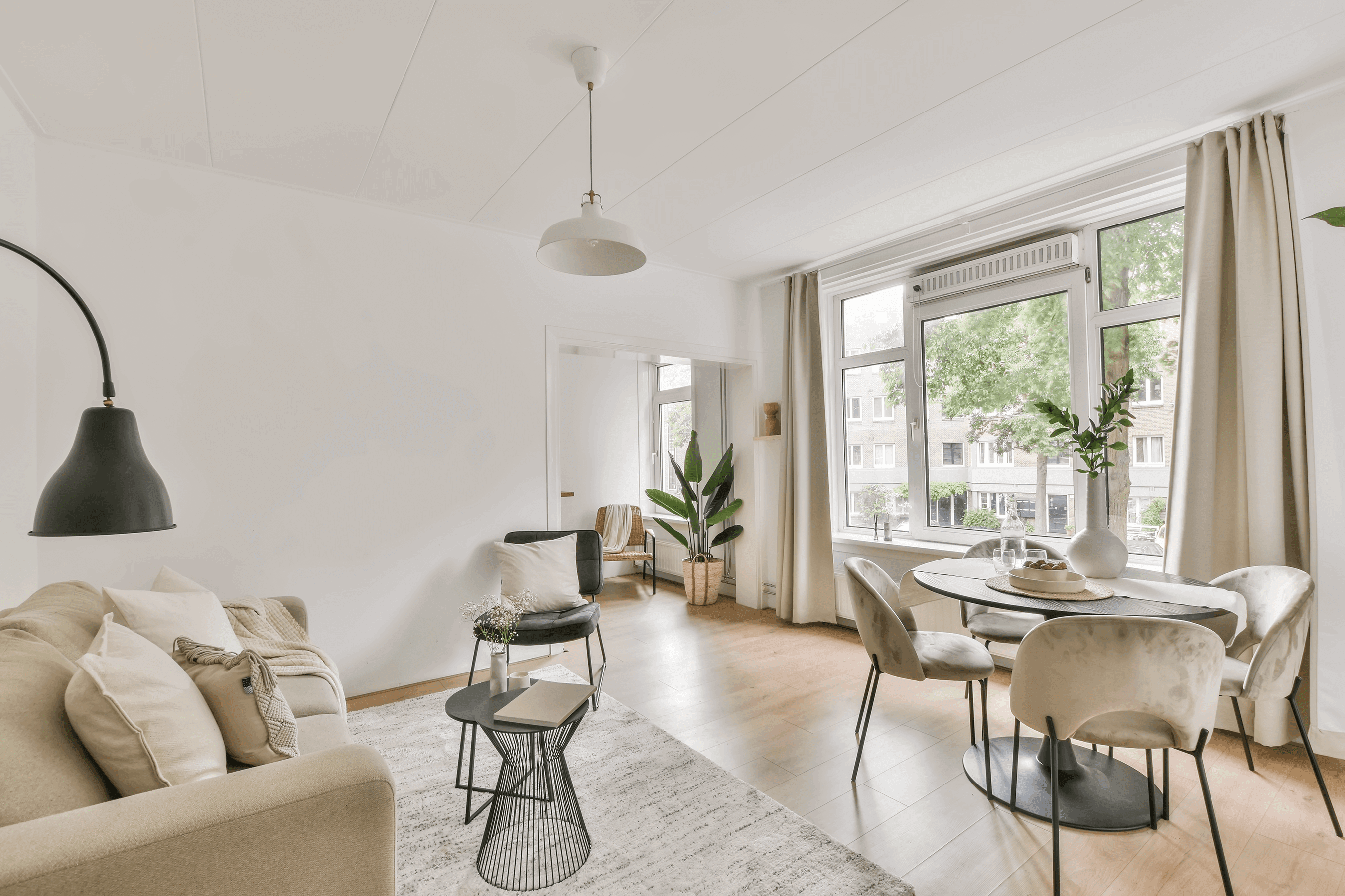 Helles, skandinavisch eingerichtetes Wohnzimmer mit beigem Sofa, schwarzer Hängeleuchte, rundem Esstisch und großen Fenstern mit Blick ins Grüne.