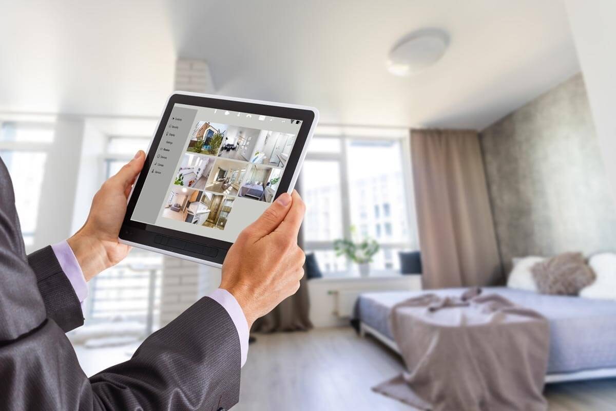 Mann hält Tablet mit Bilderübersicht einer Wohnung mit beiden Händen hoch, während im Hintergrund ein Schlafzimmer und durch die Fenster benachbarte Hochhäuser zu sehen sind. 