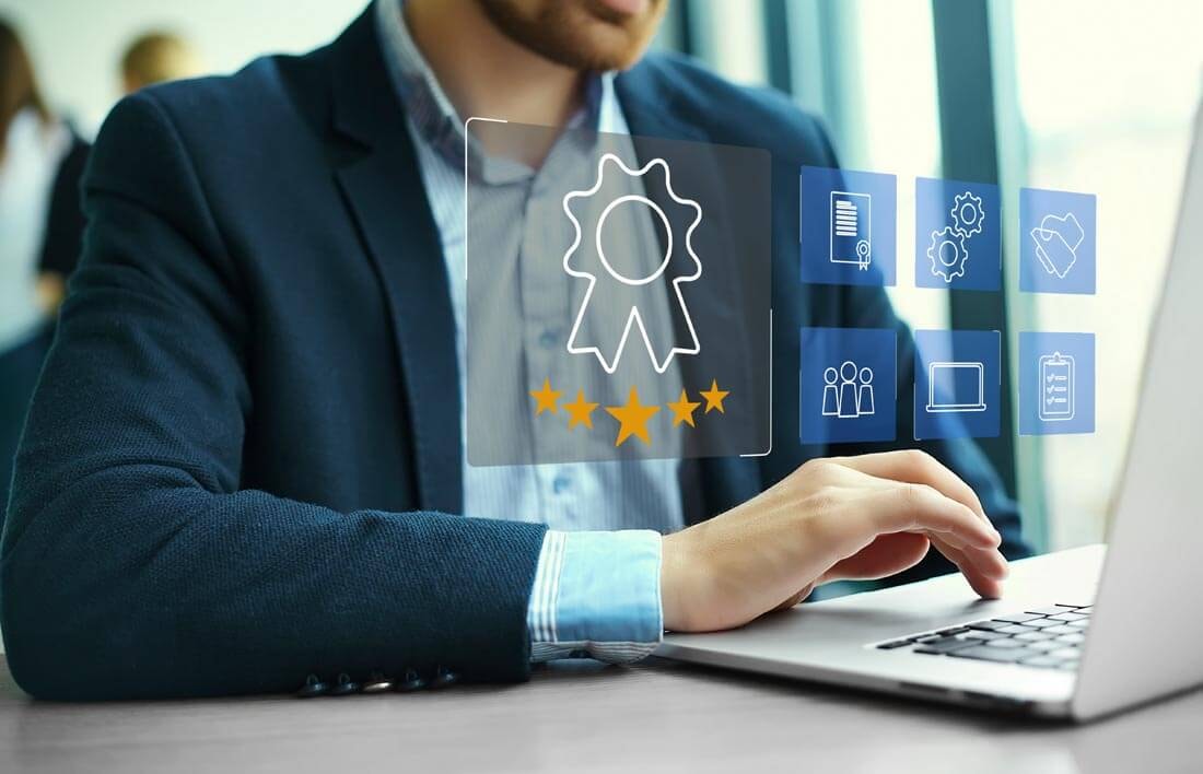 Nahaufnahme eines Mannes in Anzug der vor einem geöffneten Laptop sitzt mit der Hand am Touchpad, während vor ihm mehrere blaue, transparente Kacheln mit Icons in der Luft schweben.