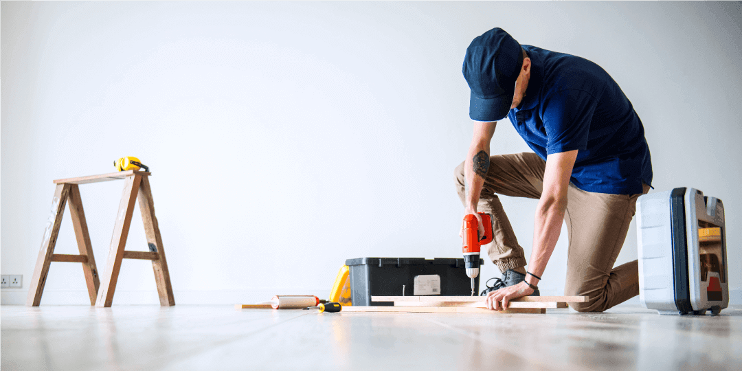 Ein Handwerker bearbeitet in einem leeren Raum Fußbodenpanele mit einer Bohrmaschine deren Koffer neben ihm liegt und einem Holzbock mit Maßband drauf, der einen Schritt weiter weg steht.