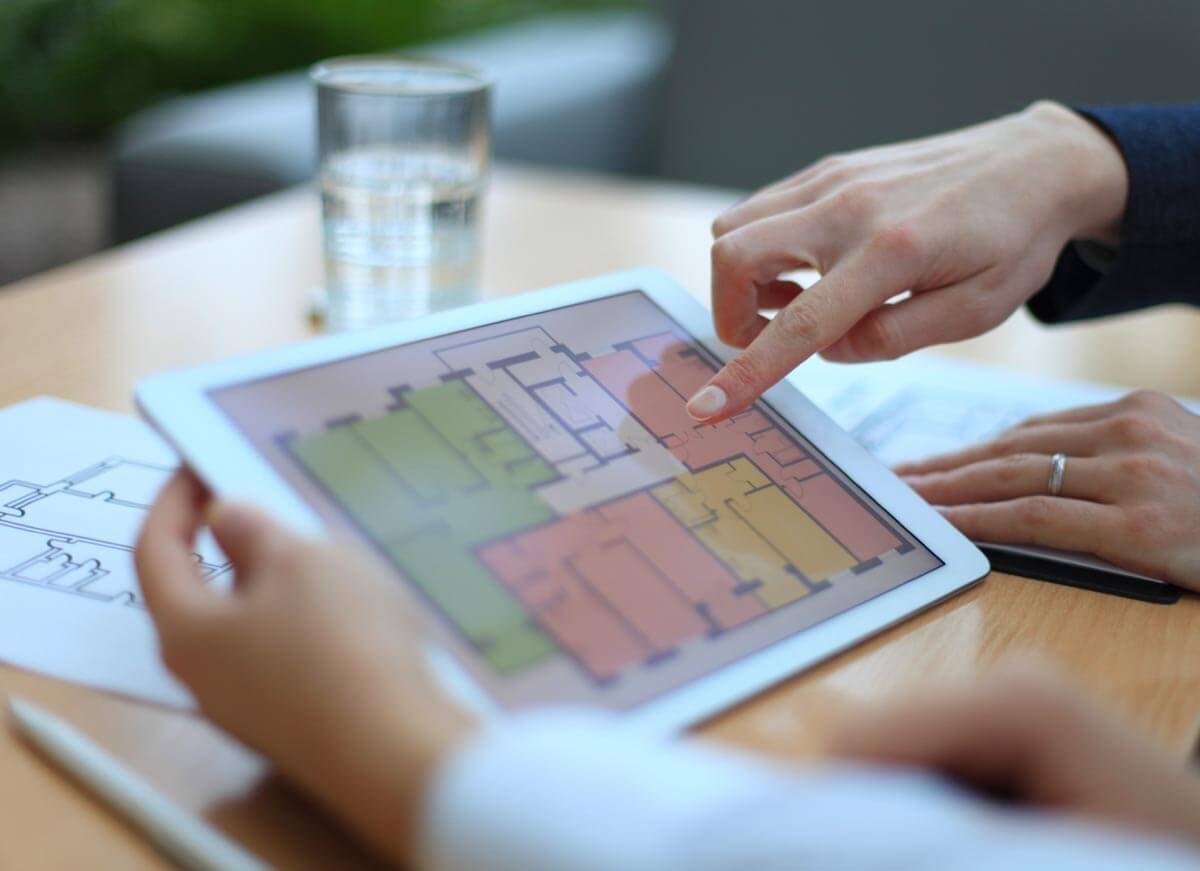 Eine Hand hält ein Tablet auf dem der Grundriss eines Mehrfamilienhauses mit Treppenhaus zu sehen ist, bei dem die einzelnen Wohneinheiten farblich getrennt sind, und eine zweite Hand zeigt mit dem Zeigefinger auf ein Detail des Grundrisses in einer der Wohneinheiten.