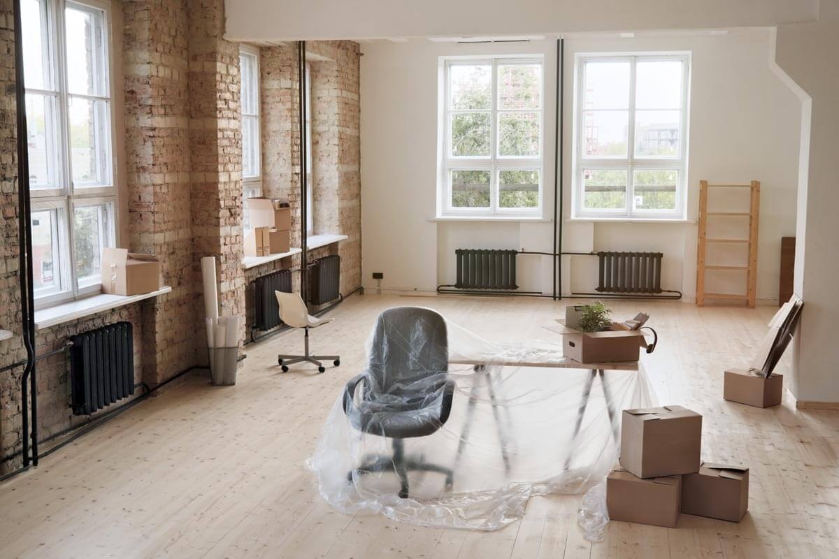 Leerer loftartiger Raum mit freiliegenden Backsteinwänden und großen Fenstern; in der Mitte steht ein in Folie eingewickelter Bürostuhl, daneben liegen Umzugskartons und ein leeres Regal steht an der Wand.