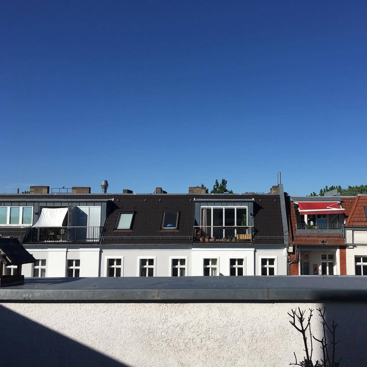 Blick von einer Dachterrasse auf eine Häuserzeile mit ausgebauten Dachgeschossen, Gauben und Balkonen vor strahlend blauem Himmel; im Vordergrund eine graue Brüstung.