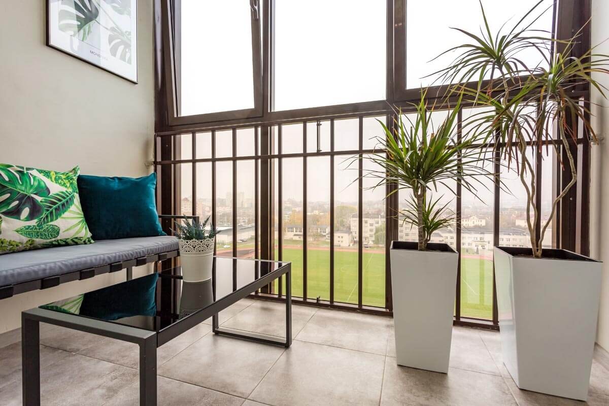 Blick aus einem verglasten Balkon über eine Sitzgruppe mit niedrigenm Tisch und Yucca-Palmen heraus auf eine Stadtlandschaft mit Hochhäusern und Grasfläche.