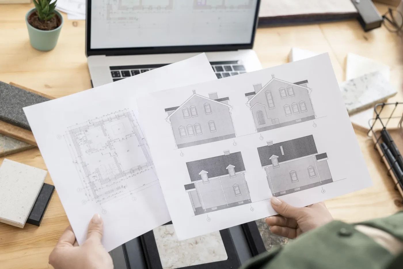 Ein Papier mit Grundriss und ein Papier mit vier Fassadenansichten eines zweigeschossigen Hauses mit Satteldach werden über einem Schreibtisch hochgehalten