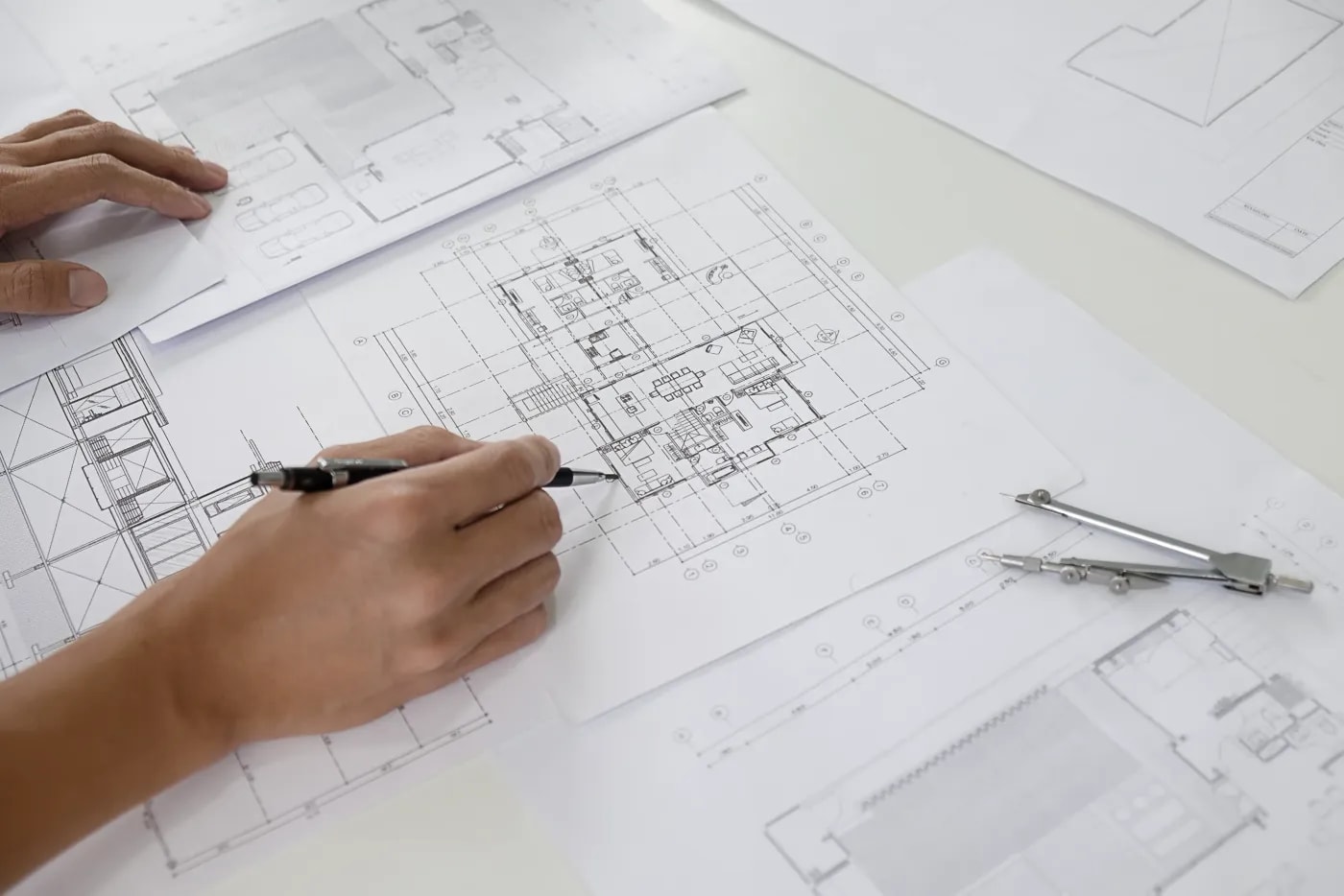 Nahaufnahme eines Bauplanes, der auf einem Tisch mit mehreren Plänen liegt und zwei Hände von denen eine einen Stift hält und ein Detail auf dem Plan nachzieht.