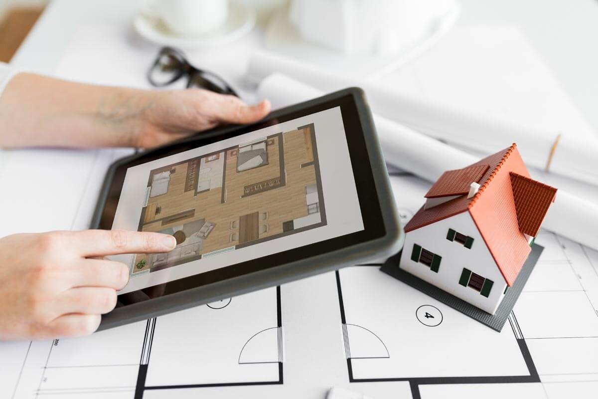 Eine Person sitzt an einem Tisch, auf dem ein Grundrissausgebreitet ist, mit Tablet in den Händen auf dem ein moderner Grundriss zu sehen ist mit einer kleinen Hausminiatur vor sich, während ihre Hand auf ein Detail im Tablet deutet.