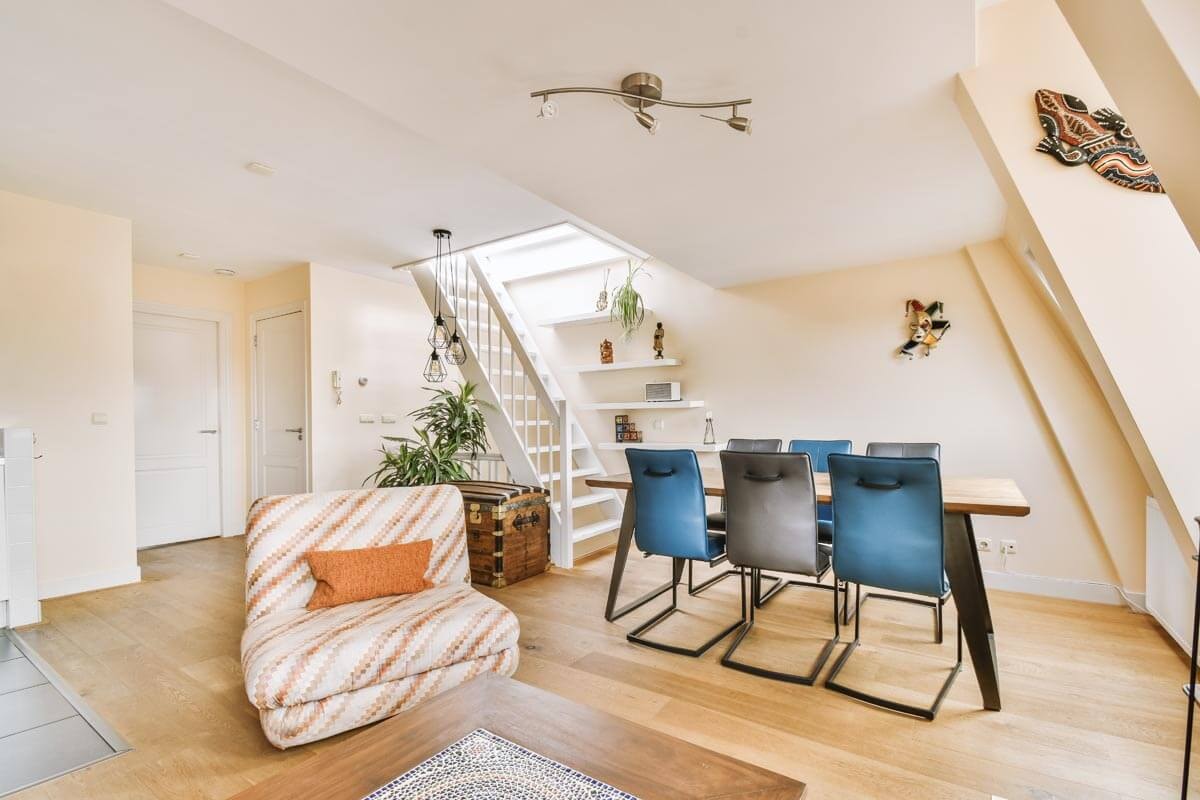 Mansardenwohnung mit hellem Parkett und charakteristischer Dachschräge rechts; Essbereich mit blauem Lederstuhl und Holztisch, links ein gemusterter Sessel mit orangem Kissen, im Hintergrund eine weiße Holztreppe nach oben mit Geländer.