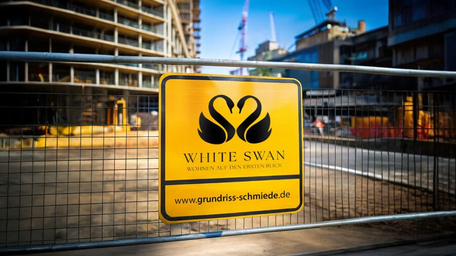 Nahaufnahme eines Teils des Bauzauns vor der Baustelle mit einem gelben Schild mit dem Logo und der Aufschrift 'White Swan' und der Webadresse der Grundriss-Schmiede.