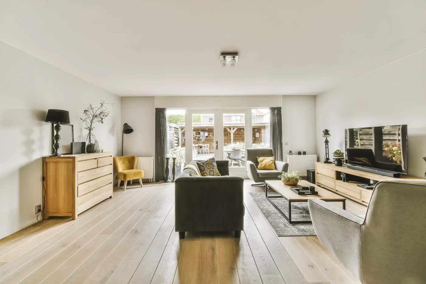Helles, langgestrecktes Wohnzimmer mit Holzfußboden und Fenster mit Terrassentüren auf der Stirnseite, durch die ein Garten mit Gartenhäuschen erkennbar ist.