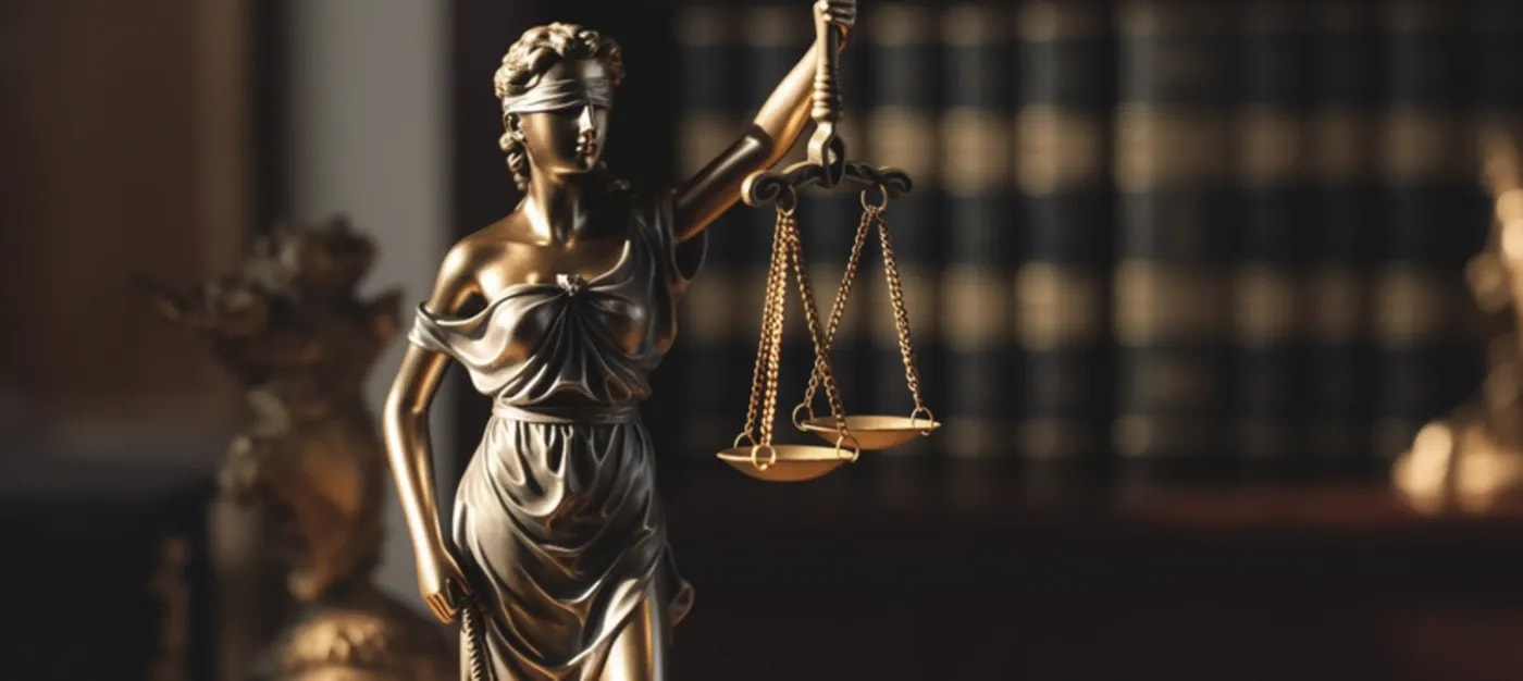 Die blinde Justitia mit Augenbinde und Waage in der Hand als Bronze-Statuette in einem Arbeitszimmer und einigen großen Büchern im Hintergrund.