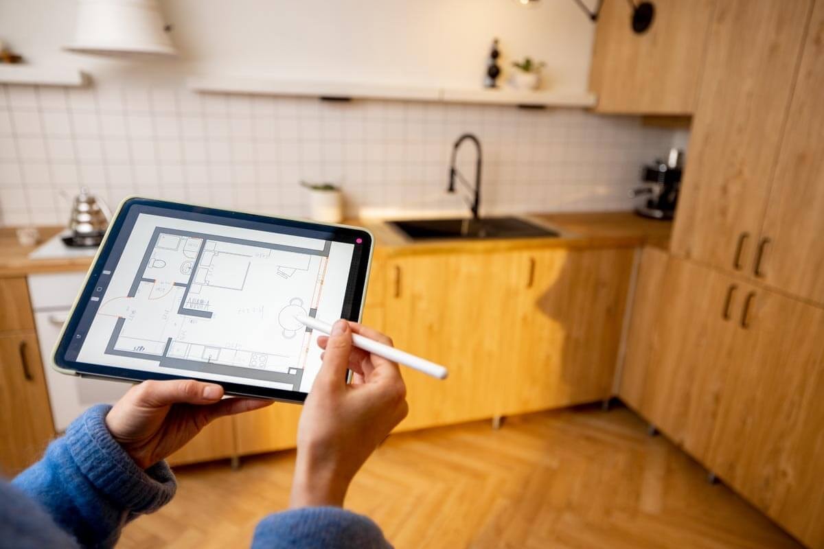 Eine Person hält ein Tablet hoch in einer Küche mit Kiefernholzschränken und Parkett.