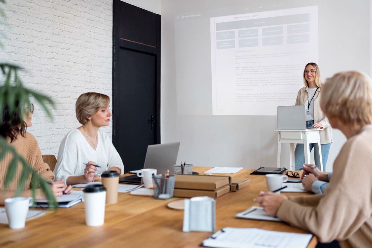 Mehrere Frauen sitzen an einem vollgestellten Holztisch bei einem Meeting, bei dem eine Frau eine Powerpoint-Präsentation hält.